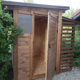 Drewniana szopa ogrodowa z otwartymi drzwiami, widoczne wnętrze z płyt OSB, obok ażurowa ściana z listew, brązowe wykończenia dachu.