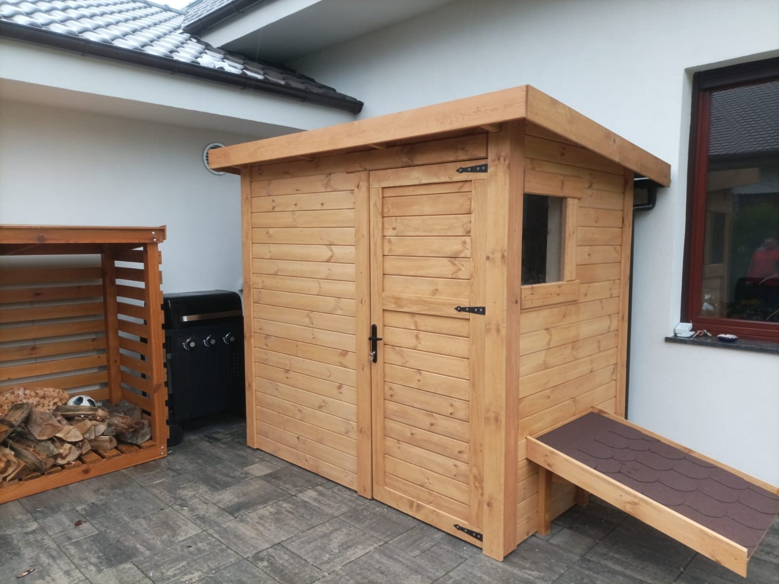 Drewniana szopa narzędziowa z oknem i małą rampą zadaszoną papą, stojąca na szarym bruku obok grilla i drewnianej konstrukcji na drewno opałowe.