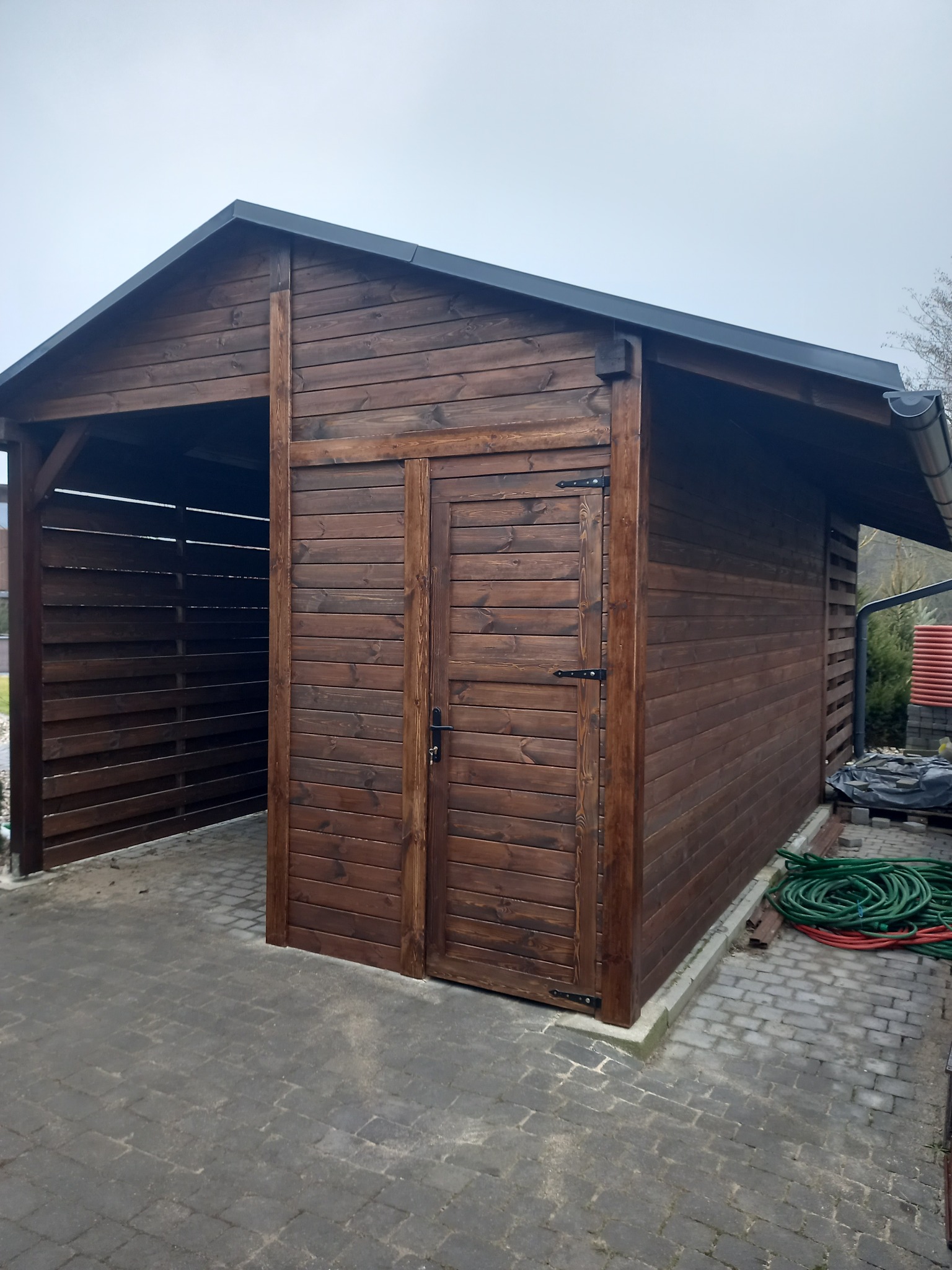 Drewniana konstrukcja z zamykaną częścią i zadaszeniem, pomalowana na ciemny brąz, widoczna z boku na tle brukowanej nawierzchni.