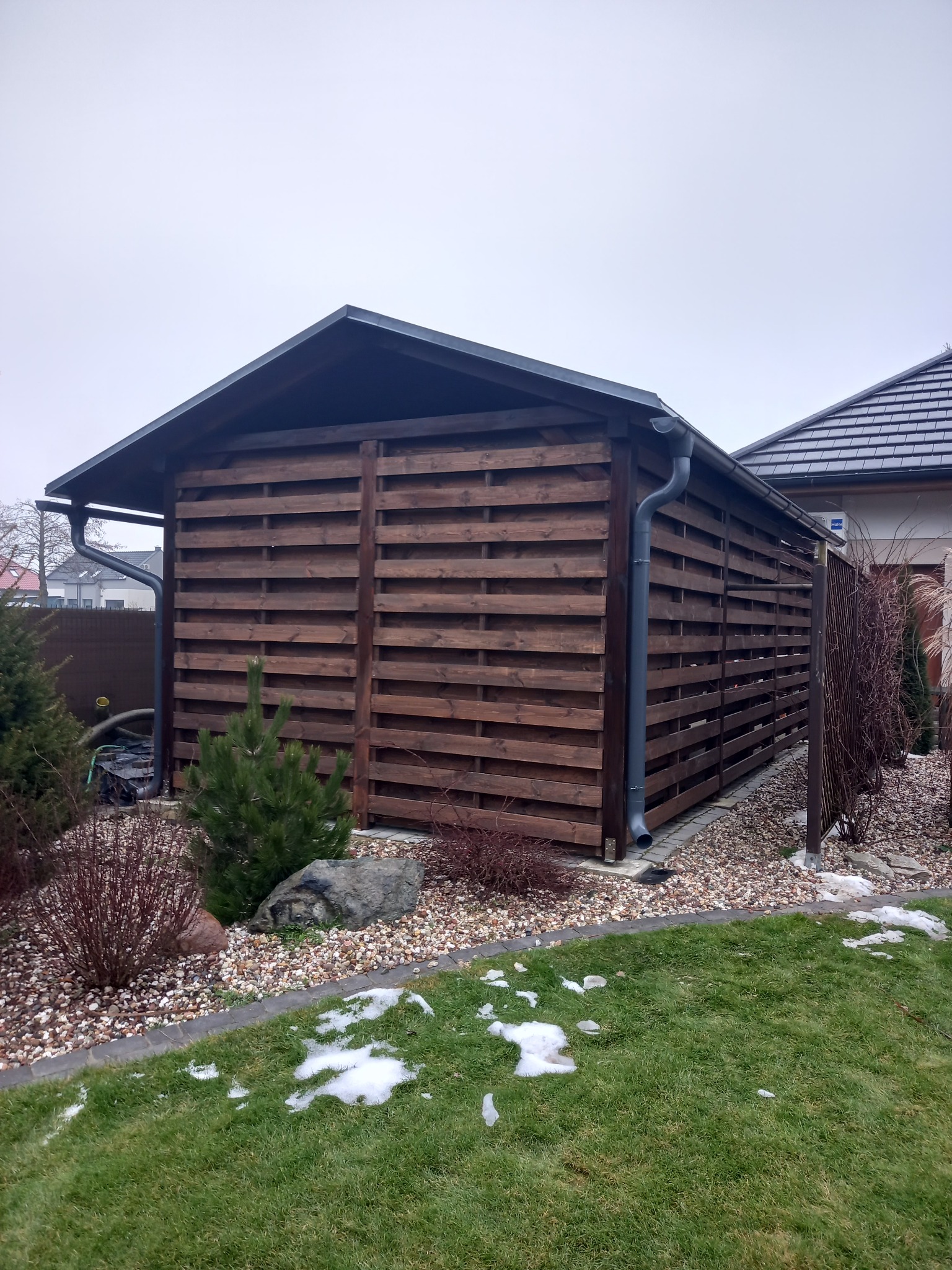 Zadaszona konstrukcja z drewnianych listew w ogrodzie, z rynną odprowadzającą wodę, otoczona kamieniami i zielenią, z fragmentami śniegu na trawie.