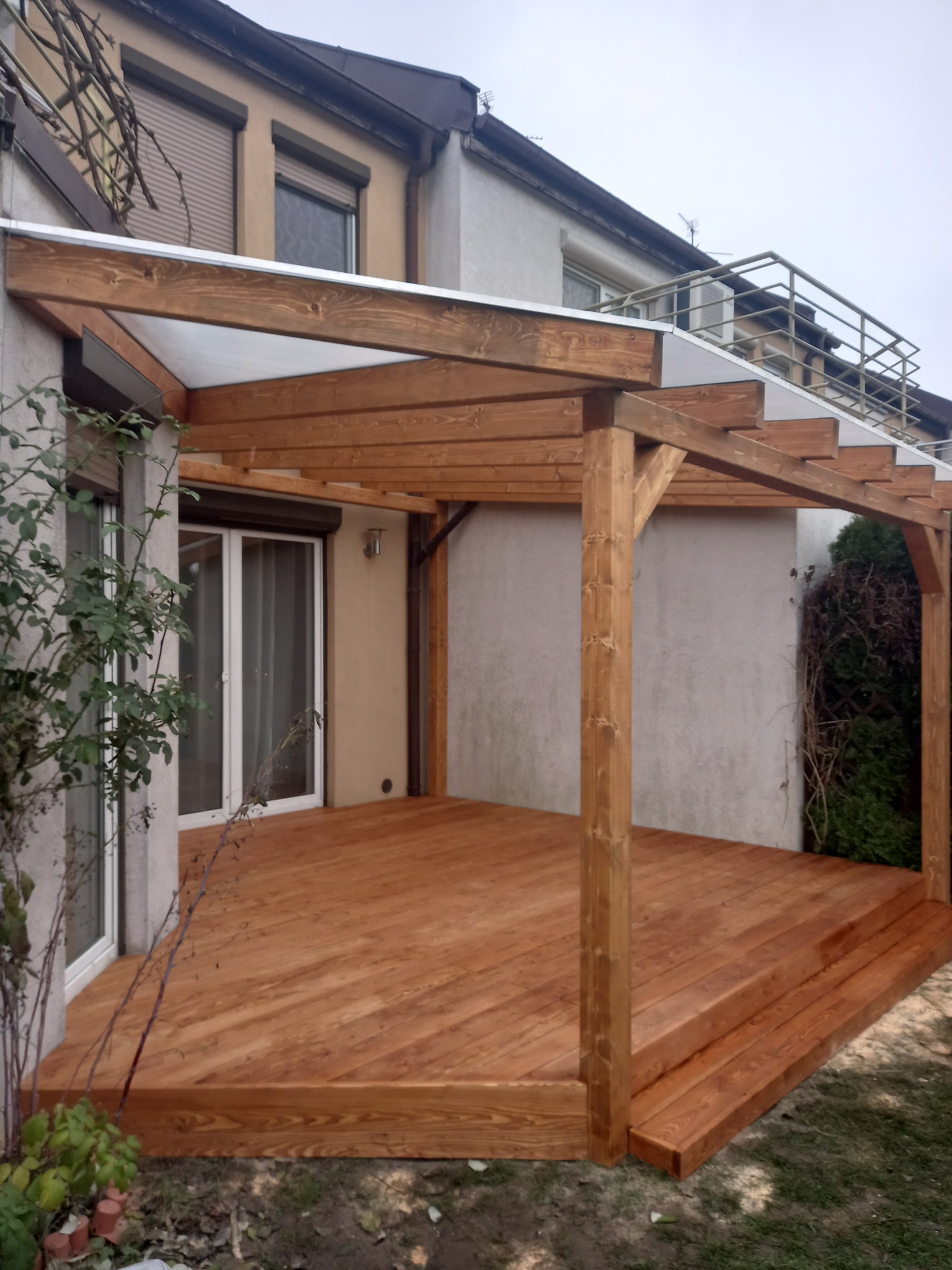 Drewniany taras przylegający do domu z zadaszeniem z przezroczystego tworzywa, widoczne dwustopniowe schody prowadzące na taras.