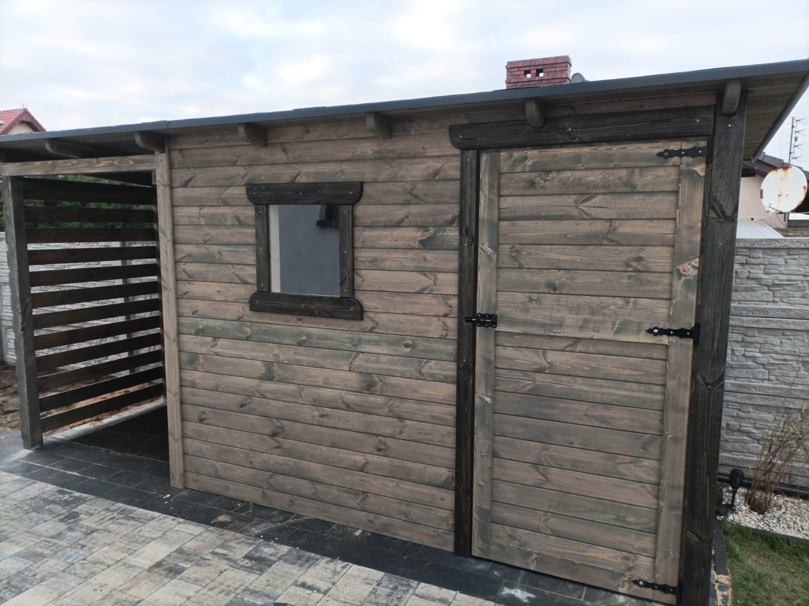 Drewniana altana z małym oknem i drzwiami, pomalowana na szaro-brązowy kolor, z czarnymi okuciami, widoczna z boku, z zadaszeniem i elementami ażurowymi po lewej stronie, na kostce brukowej.