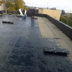 Prace dekarskie na płaskim dachu: ekipa robotników rozprowadza masę bitumiczną, widoczne rolki papy i przygotowane podłoże, budynek w tle.