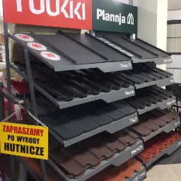 Ekspozycja różnych modeli i kolorów pokryć dachowych na metalowym stojaku w sklepie, z widocznymi nazwami producentów Ruukki i Plannja oraz tabliczką 'Zapraszamy po wyroby hutnicze'.