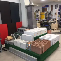Ekspozycja różnorodnych materiałów budowlanych w sklepie: bloczki betonowe, pustaki ceramiczne, cegły, elementy kominowe. Widoczny aplikator do kleju na jednym z elementów. W tle biuro obsługi...