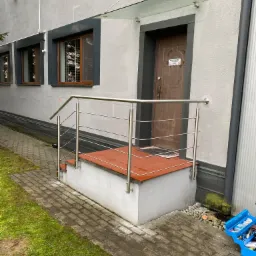 Zewnętrzne schody wejściowe z terakotowymi płytkami i stalową poręczą z poziomymi wypełnieniami, osłonięte szklanym daszkiem, na tle szarej elewacji budynku z oknami.