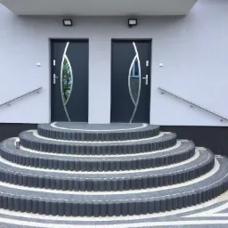 Półokrągłe schody z szarej kostki brukowej prowadzące do dwóch nowoczesnych, grafitowych drzwi z elementami ze stali nierdzewnej i szklanymi wstawkami, z zamontowanymi poręczami po bokach.