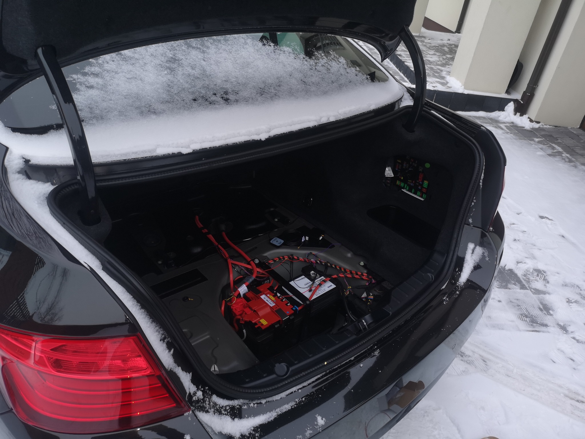 Otwarty bagażnik czarnego samochodu, widoczna instalacja elektryczna i akumulator, na zewnątrz śnieg.