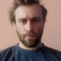 Portret zamyślonego mężczyzny z brodą i rozczochranymi włosami, ubrany w ciemny t-shirt na tle jednolitej ściany.