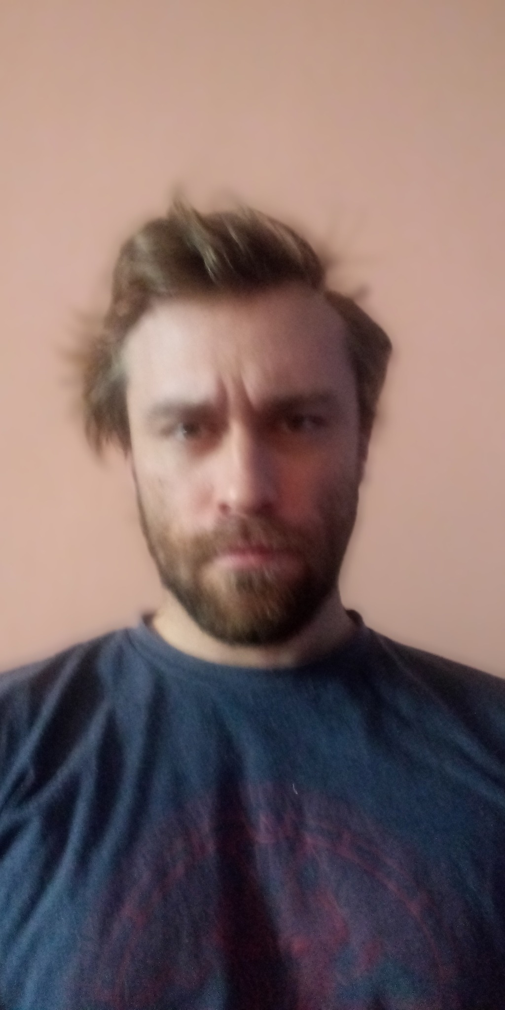 Portret zamyślonego mężczyzny z brodą i rozczochranymi włosami, ubrany w ciemny t-shirt na tle jednolitej ściany.