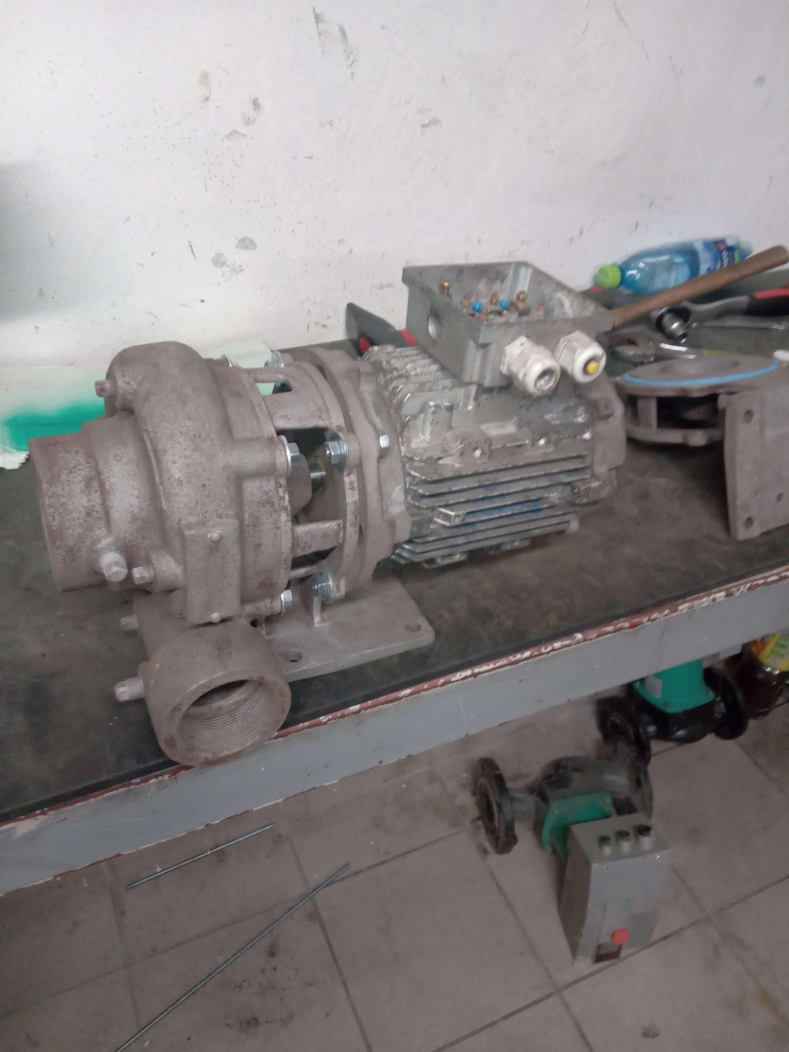 Zdemontowany, zardzewiały silnik pompy obiegowej na warsztatowym stole, z widocznym uzwojeniem, skrzynką elektryczną i elementami montażowymi; obok leży butelka wody.