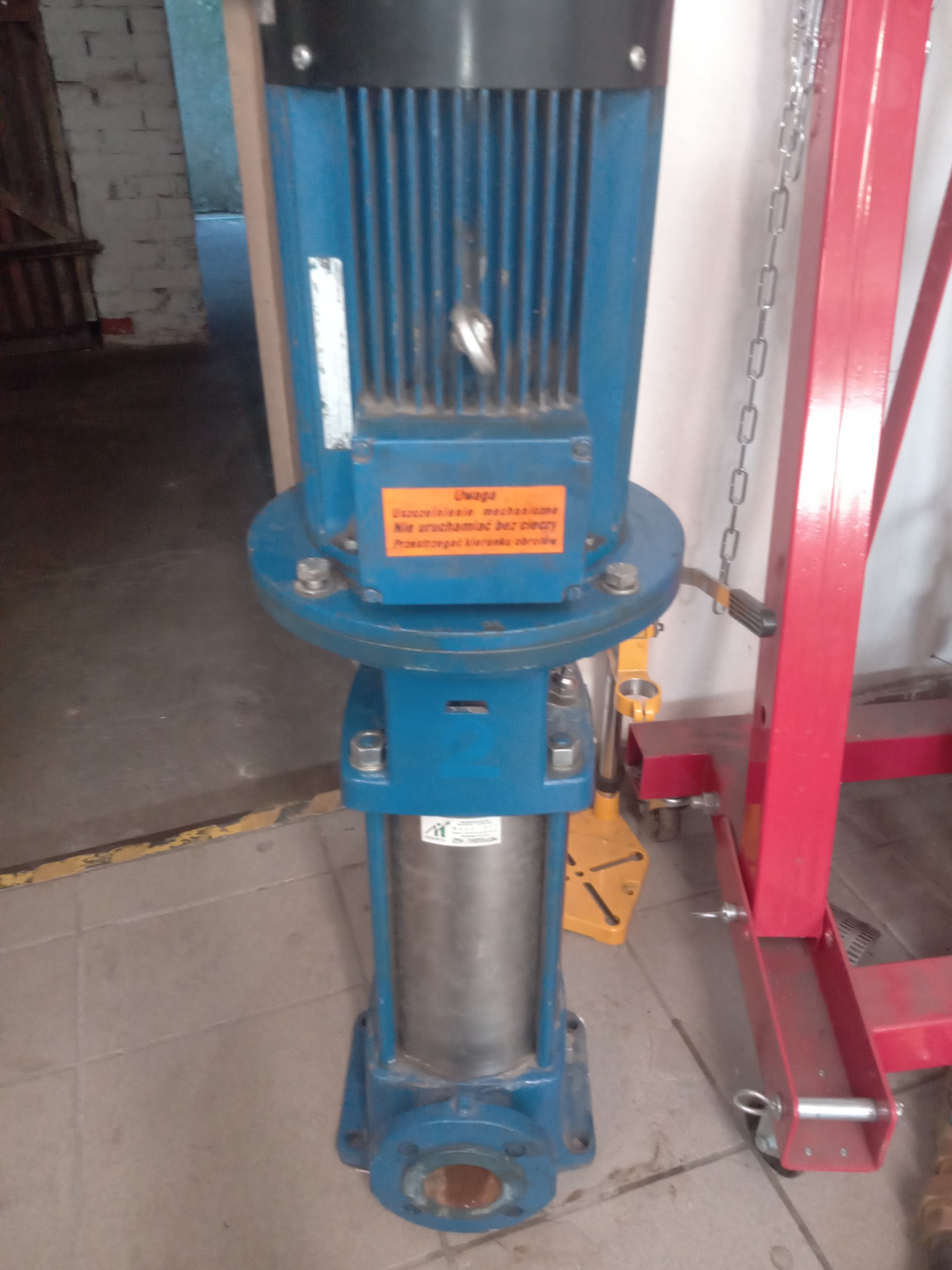 Pionowa pompa wielostopniowa, niebieska obudowa z silnikiem u góry, pomarańczowa naklejka ostrzegawcza, widoczna część urządzenia do podnoszenia silników w kolorze czerwonym.