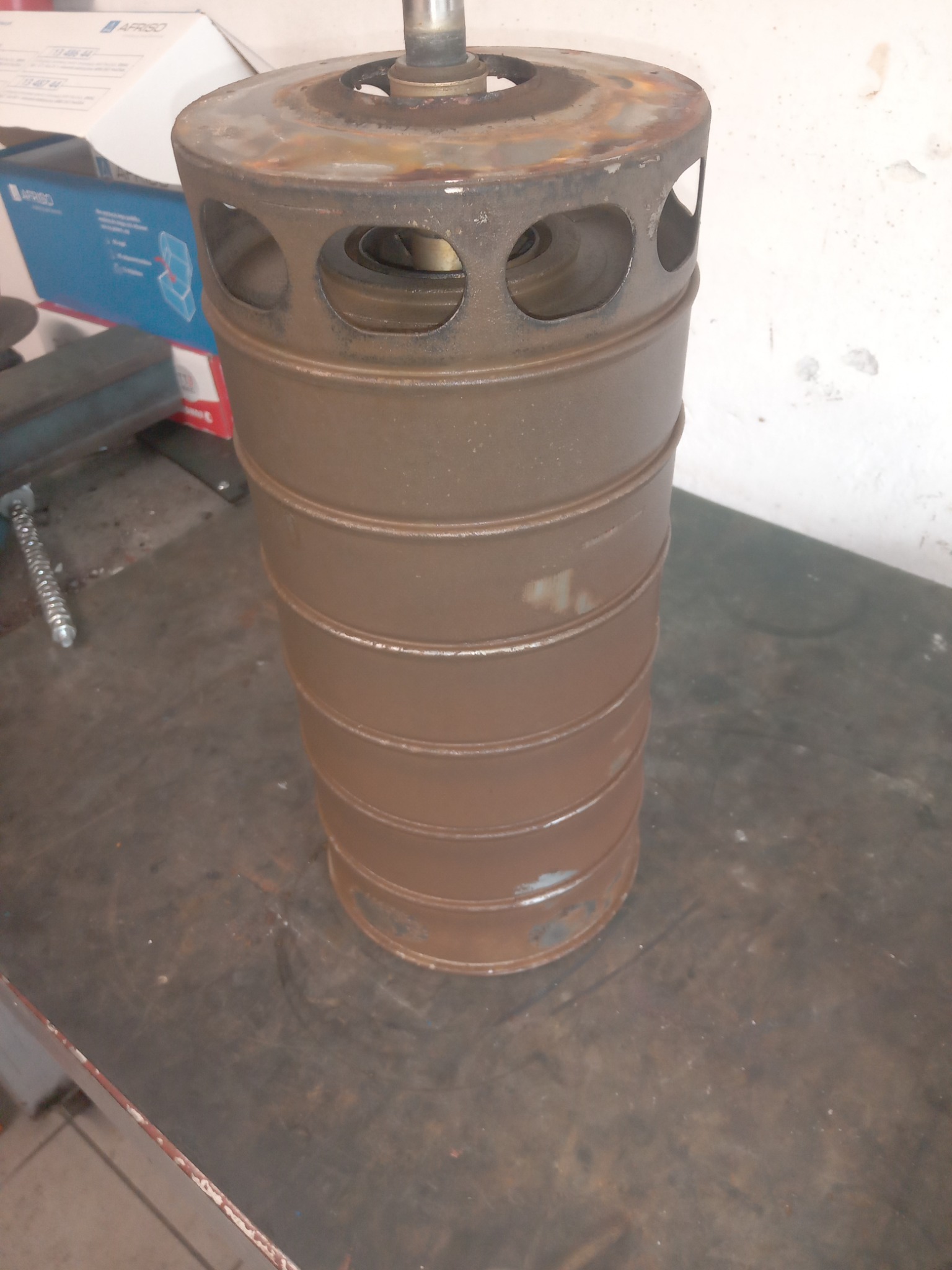 Metalowy, cylindryczny element pieca z otworami wentylacyjnymi, widoczne ślady rdzy i zużycia, ustawiony na brudnym blacie warsztatowym, w tle pudełko AFRISO.