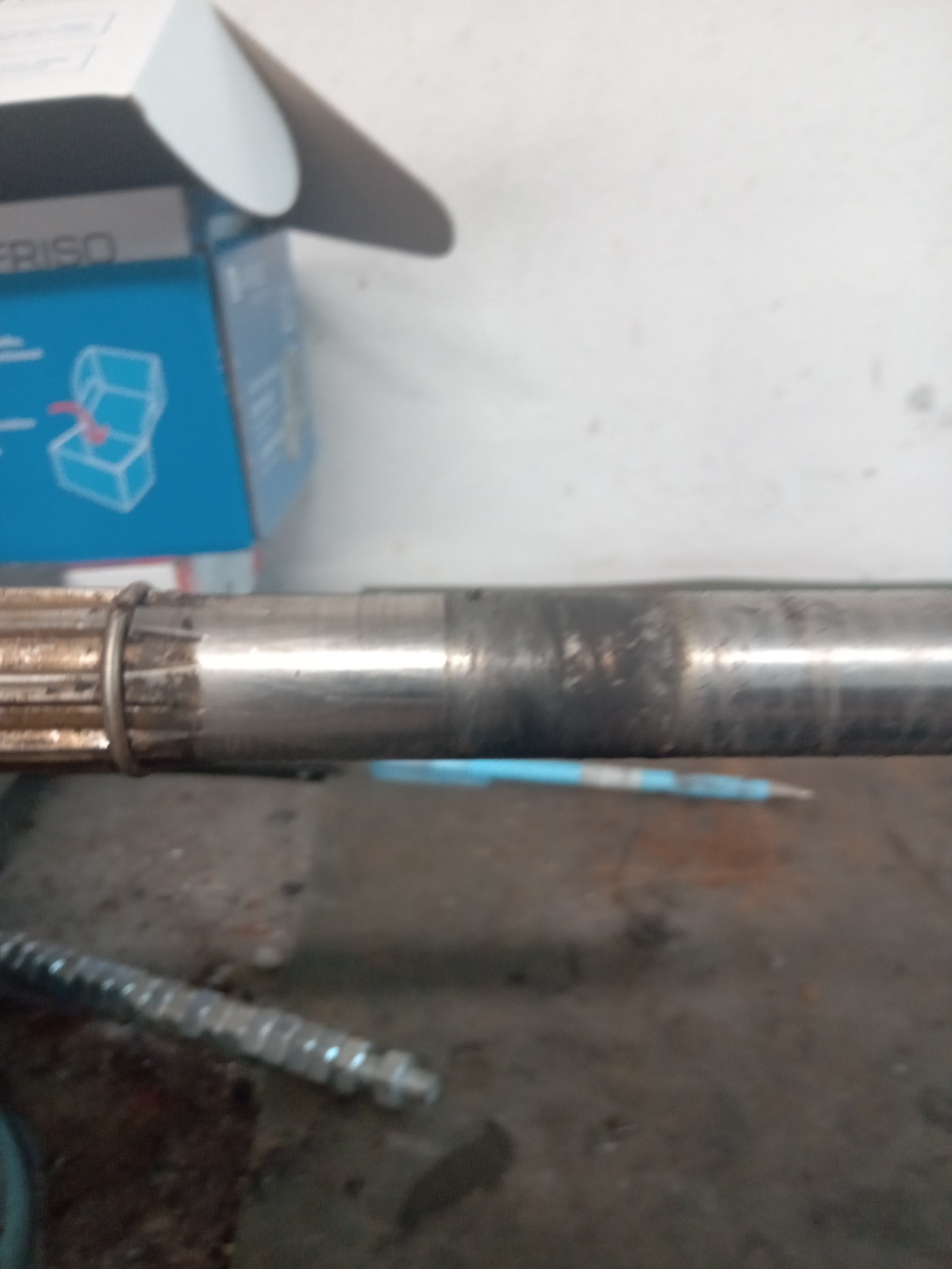 Metalowy wał z zębatką, widoczne ślady użytkowania i korozji, obok niebieski ołówek i pudełko z instrukcją montażu.