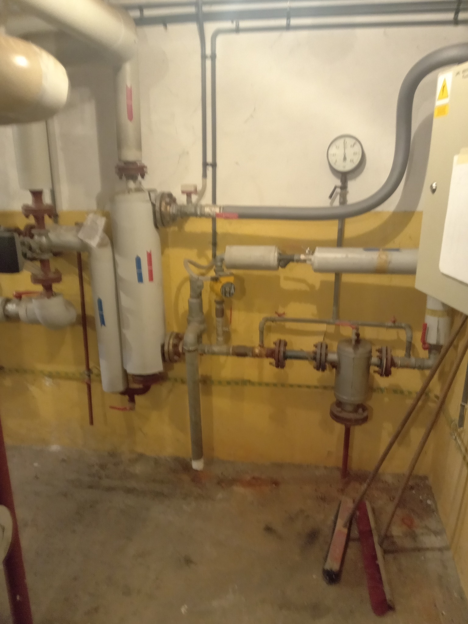 Węzeł cieplny w piwnicy z licznymi rurami, filtrami i manometrem, widoczne również dwie szczotki do zamiatania podłogi.