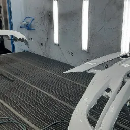 Pomalowane na biało elementy karoserii samochodowej, w tym zderzaki, suszone w kabinie lakierniczej z widocznymi lampami i kratą podłogową, czarny samochód w tle.