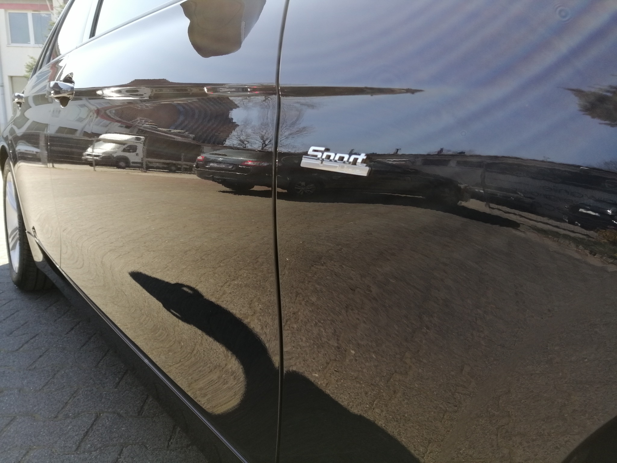 Błyszczący, czarny bok samochodu z chromowanym napisem 'Sport', odbijający otoczenie, w tym inne pojazdy i budynki.