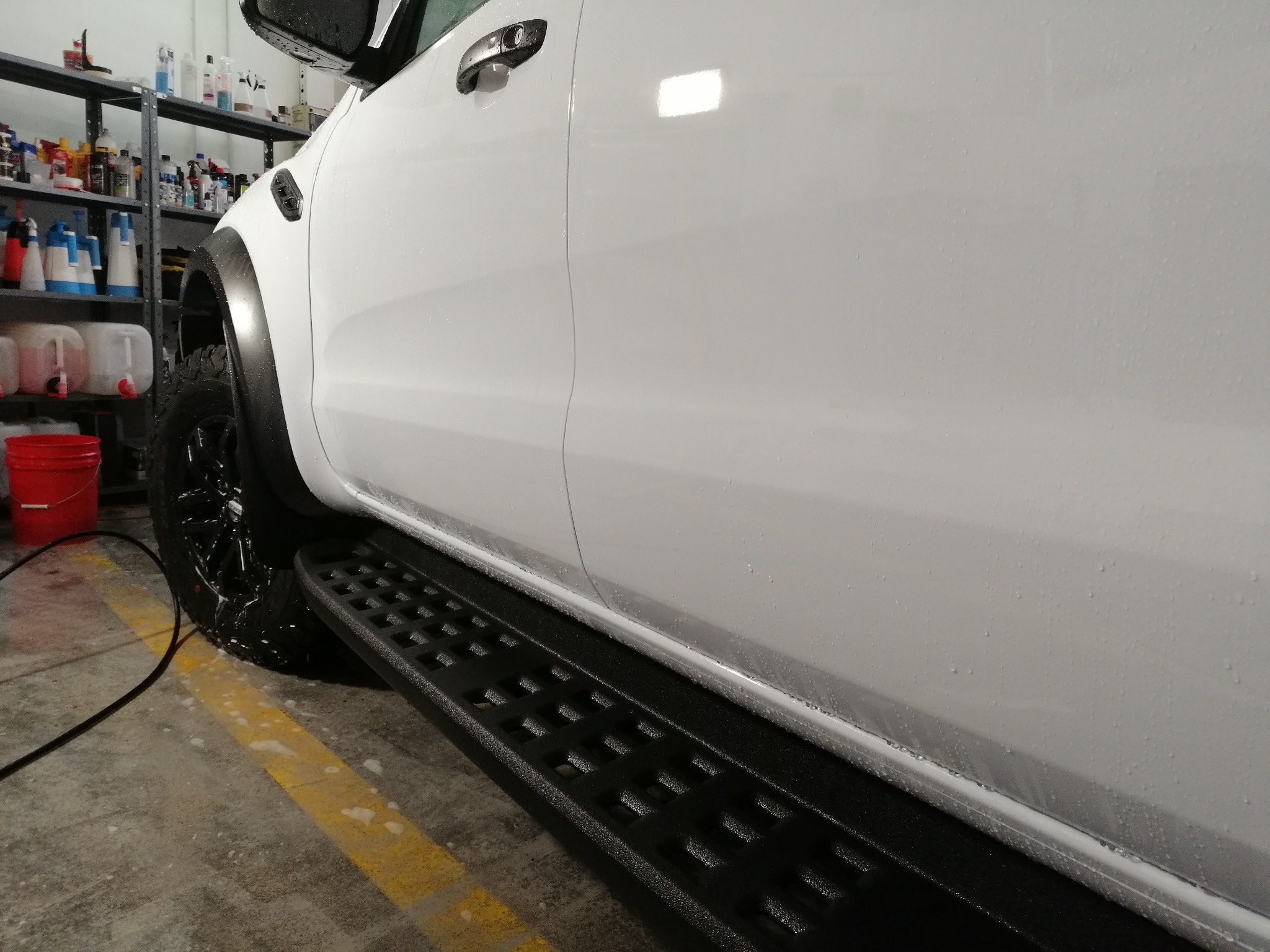 Biały samochód typu pickup, bok pojazdu pokryty kroplami wody po myciu, widoczna opona z czarną felgą i czarny stopień, w tle regał z różnymi środkami czyszczącymi.