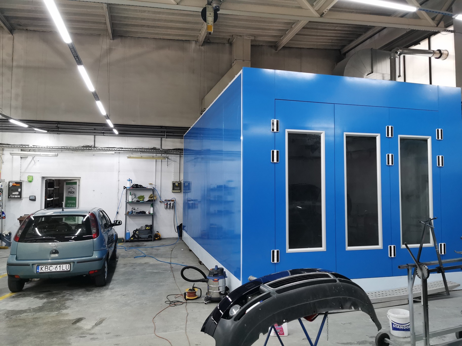 Wnętrze warsztatu z niebieską kabiną lakierniczą, szarym Oplem Corsą i czarnym zdemontowanym zderzakiem na stojaku.