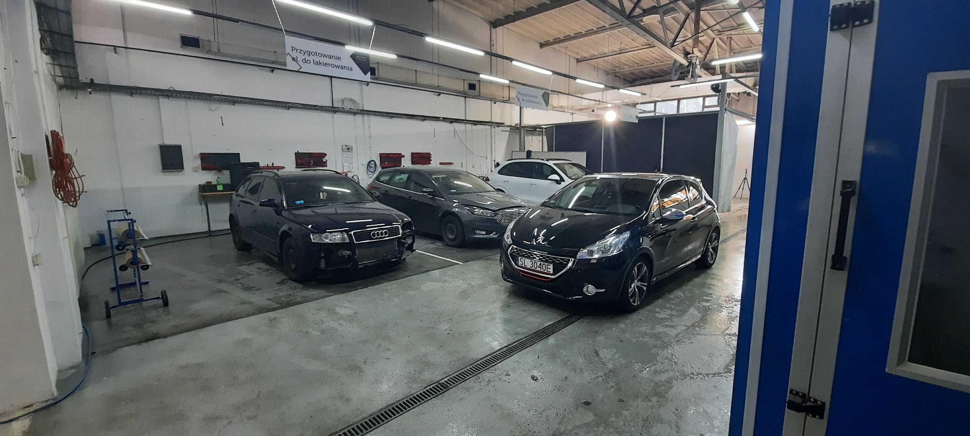 Wnętrze warsztatu samochodowego z kilkoma autami, w tym Audi A4 z widocznymi uszkodzeniami karoserii i Peugeot 208 zaparkowanymi w oczekiwaniu na naprawę, widoczny w tle boks lakierniczy.