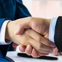 Uścisk dłoni dwóch mężczyzn w garniturach nad dokumentami i długopisem, symbolizujący zawarcie umowy.