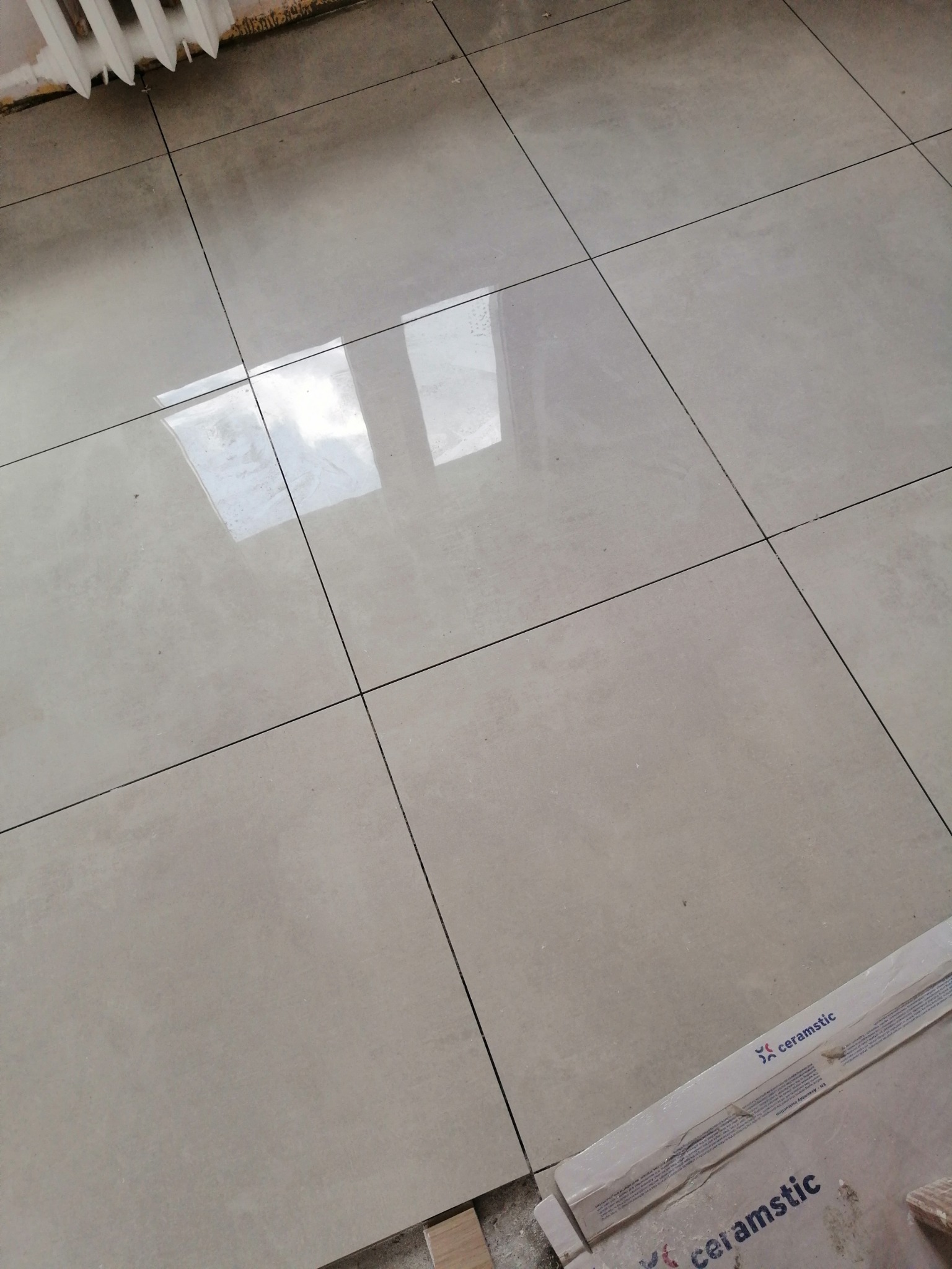 Ułożone beżowe płytki podłogowe z widocznym odbiciem okna, w rogu karton po płytkach marki Ceramstic.