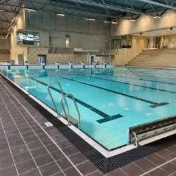 Wnętrze nowoczesnej pływalni z jasnoniebieską wodą w basenie, metalowymi drabinkami wejściowymi i stanowiskami startowymi, widok na trybuny i betonowo-stalową konstrukcję dachu.