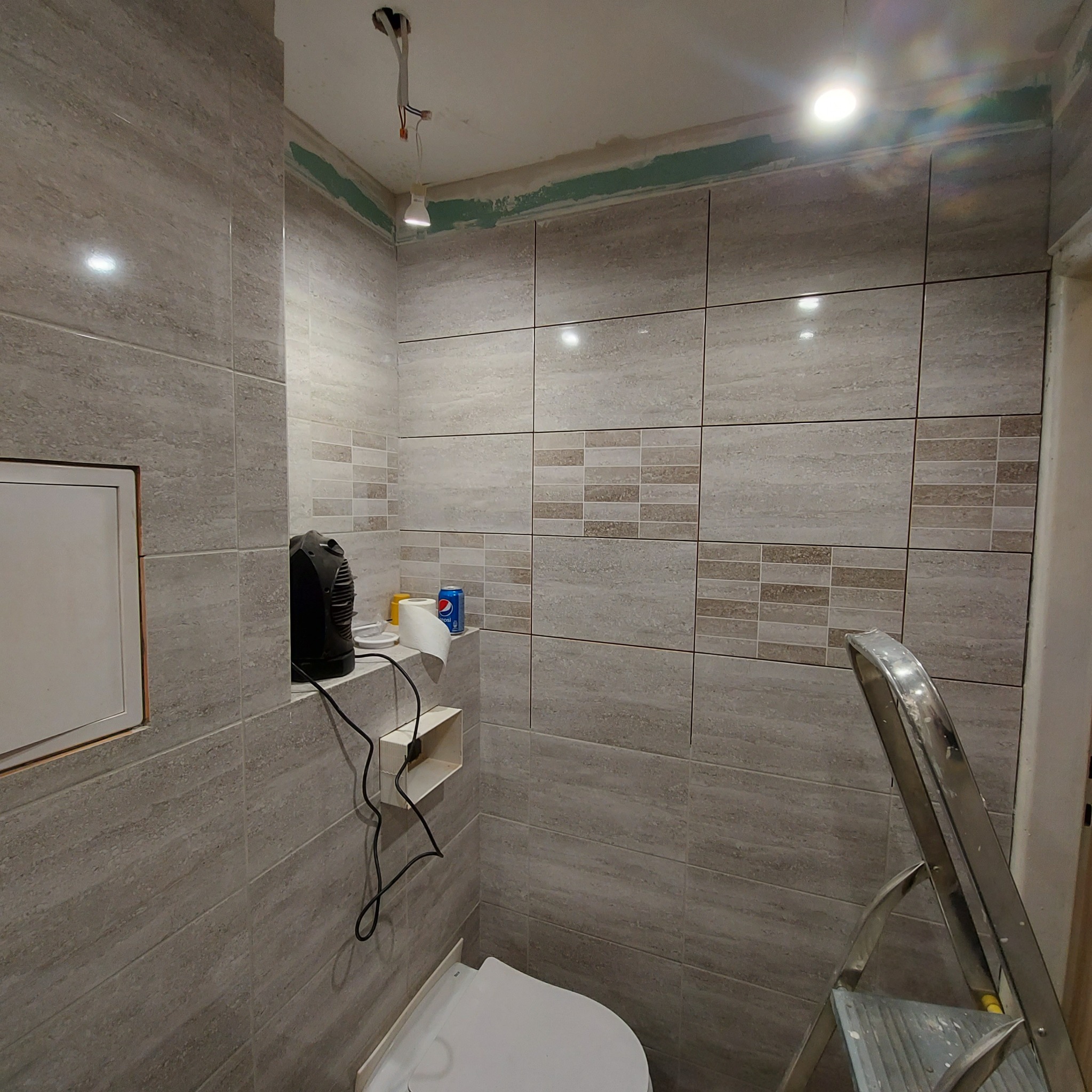 Łazienka w trakcie prac wykończeniowych: ułożone płytki imitujące kamień w odcieniach szarości i beżu, widoczna drabina aluminiowa, grzejnik elektryczny, puszka elektryczna bez osprzętu...