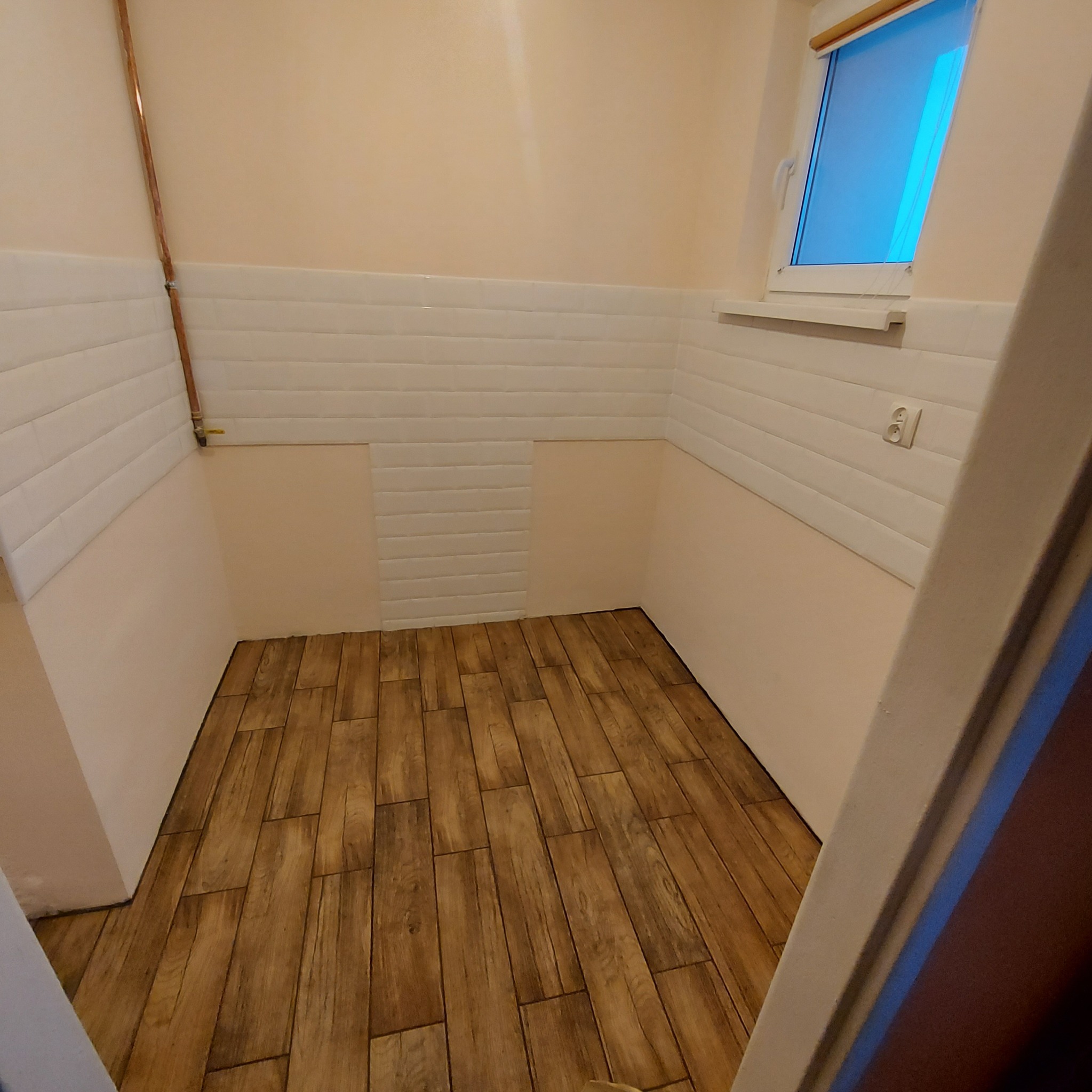 Wnętrze małej łazienki z drewnopodobną podłogą, białymi kafelkami na ścianach do połowy wysokości i beżowymi ścianami powyżej, widoczne okno z roletą.