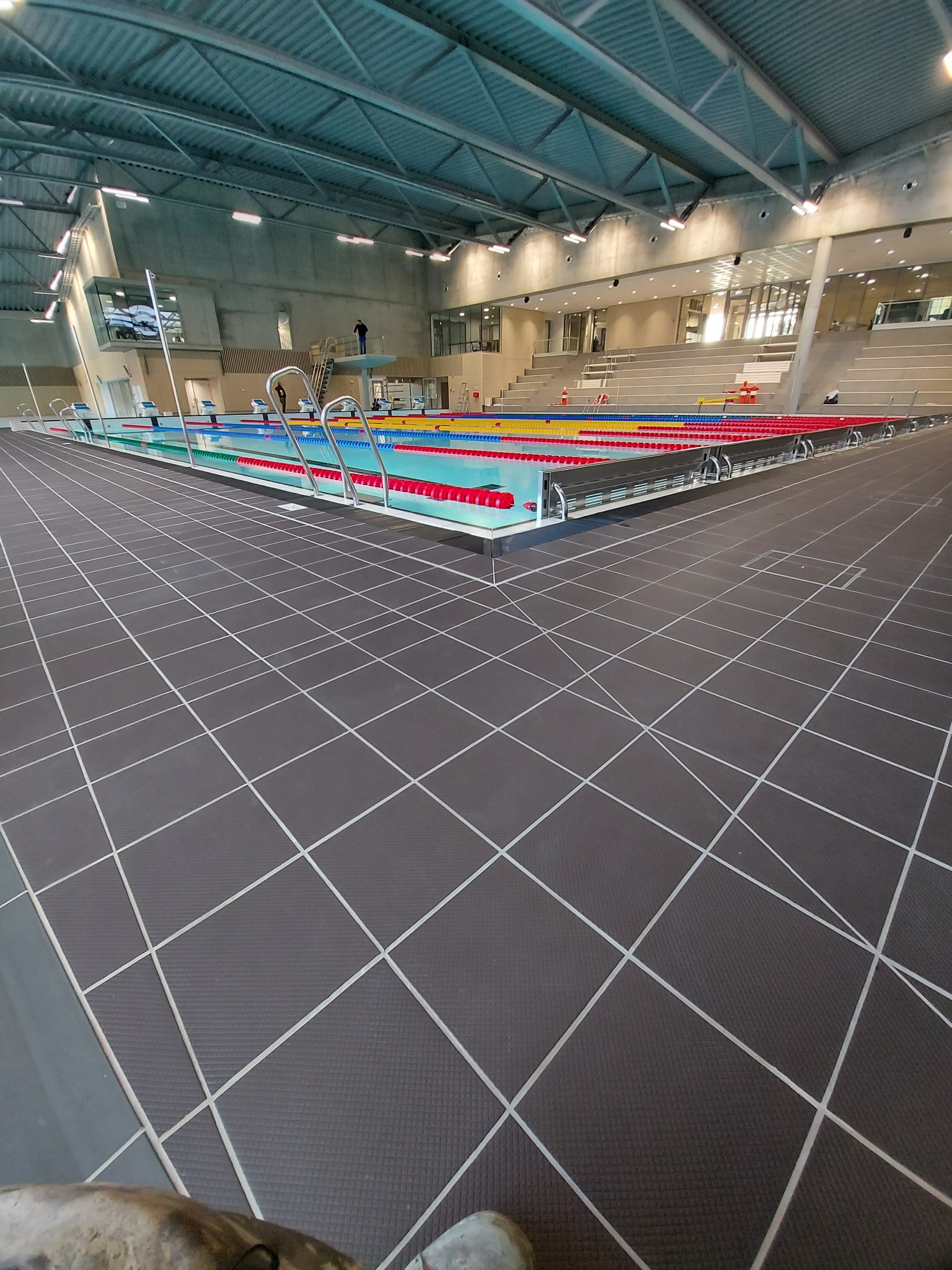Wnętrze hali basenowej z zaznaczonymi torami pływackimi, widok z perspektywy podłogi z ciemnych płytek z jasnymi liniami, w tle trybuny i wieża do skoków.