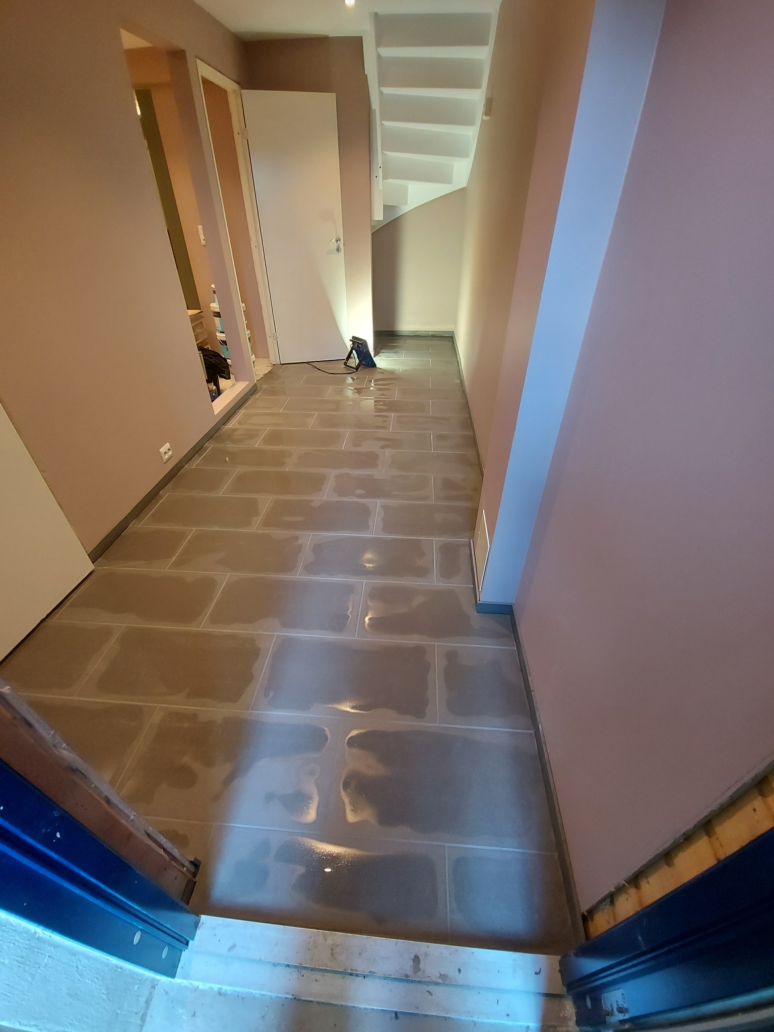 Korytarz w trakcie układania płytek podłogowych, widoczna poziomica laserowa, szare płytki prostokątne z efektem mokrej powierzchni, różowe ściany, białe schody i drzwi.