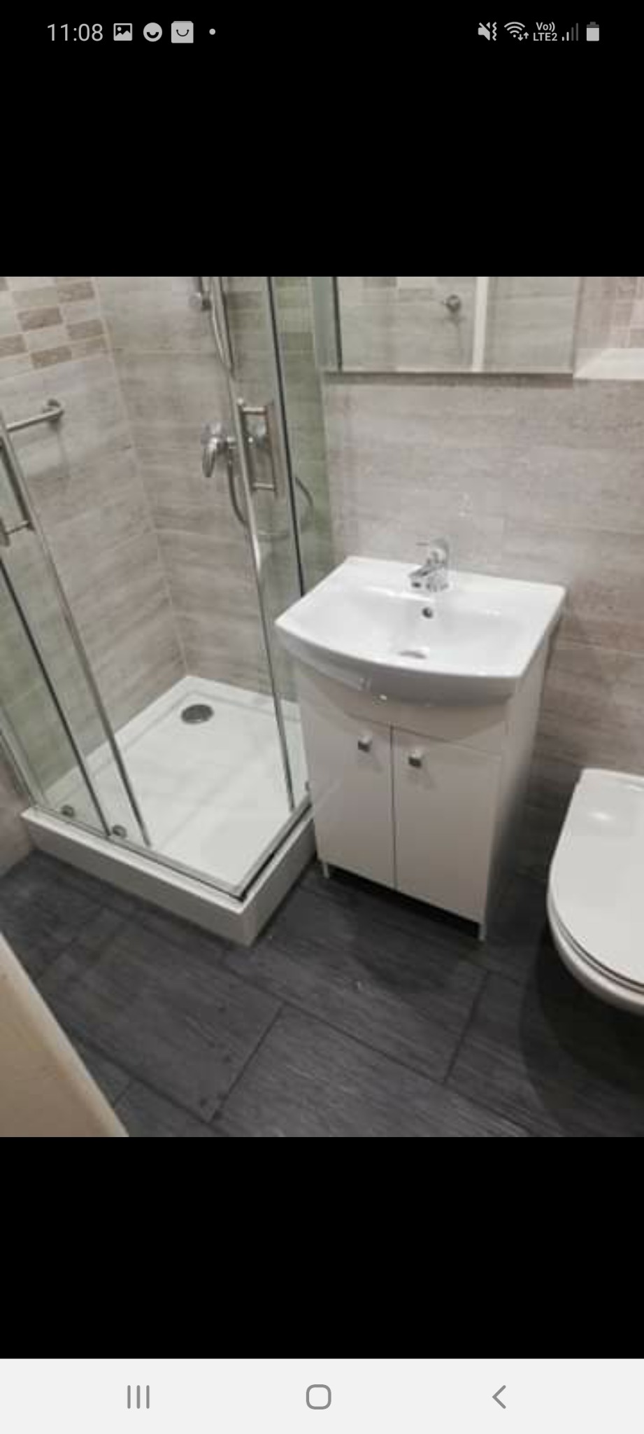 Nowoczesna łazienka z kabiną prysznicową, umywalką z szafką i toaletą, wykończona w odcieniach szarości i bieli z ciemną podłogą imitującą drewno.