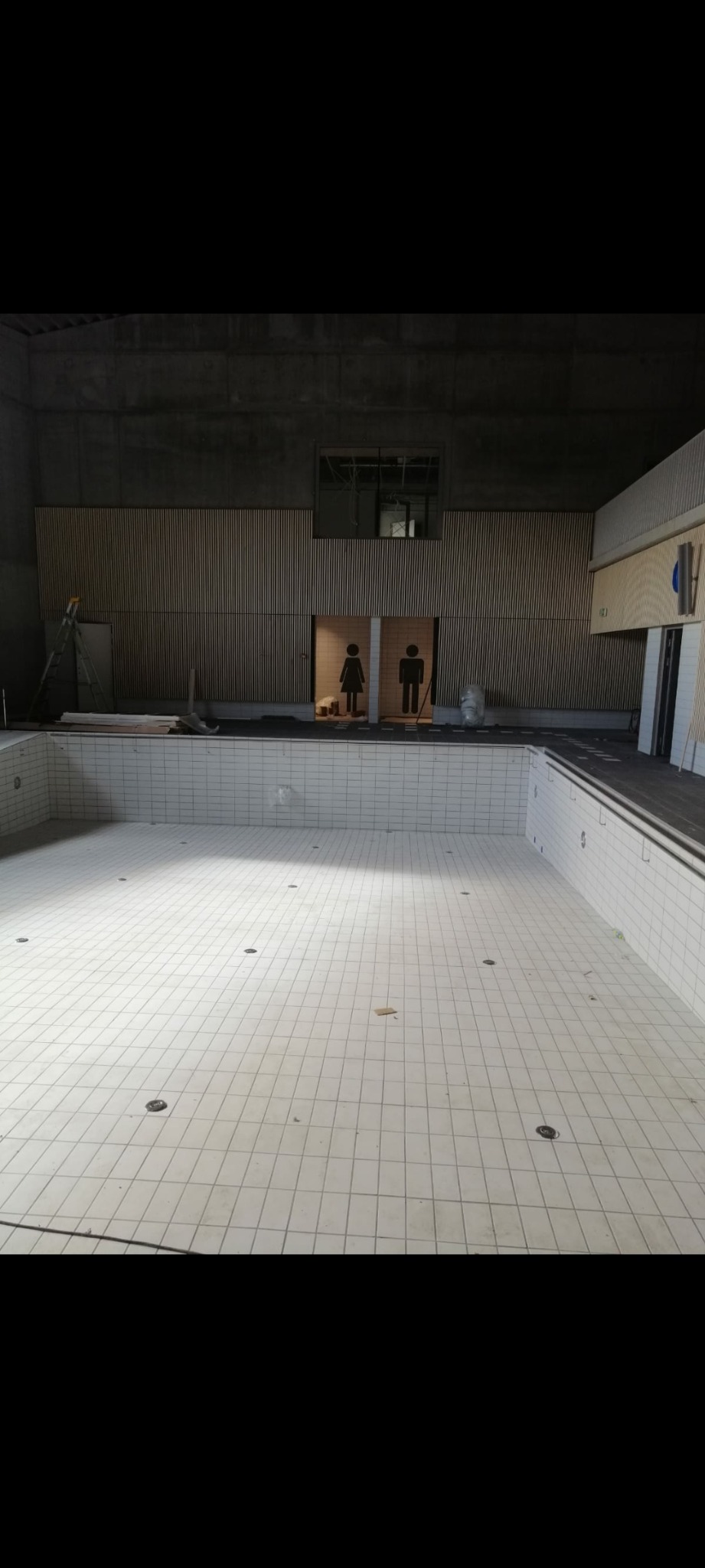 Wnętrze remontowanego basenu z białymi płytkami, widoczne wejścia do przebieralni oznaczone piktogramami kobiety i mężczyzny, drabina i materiały budowlane w tle.