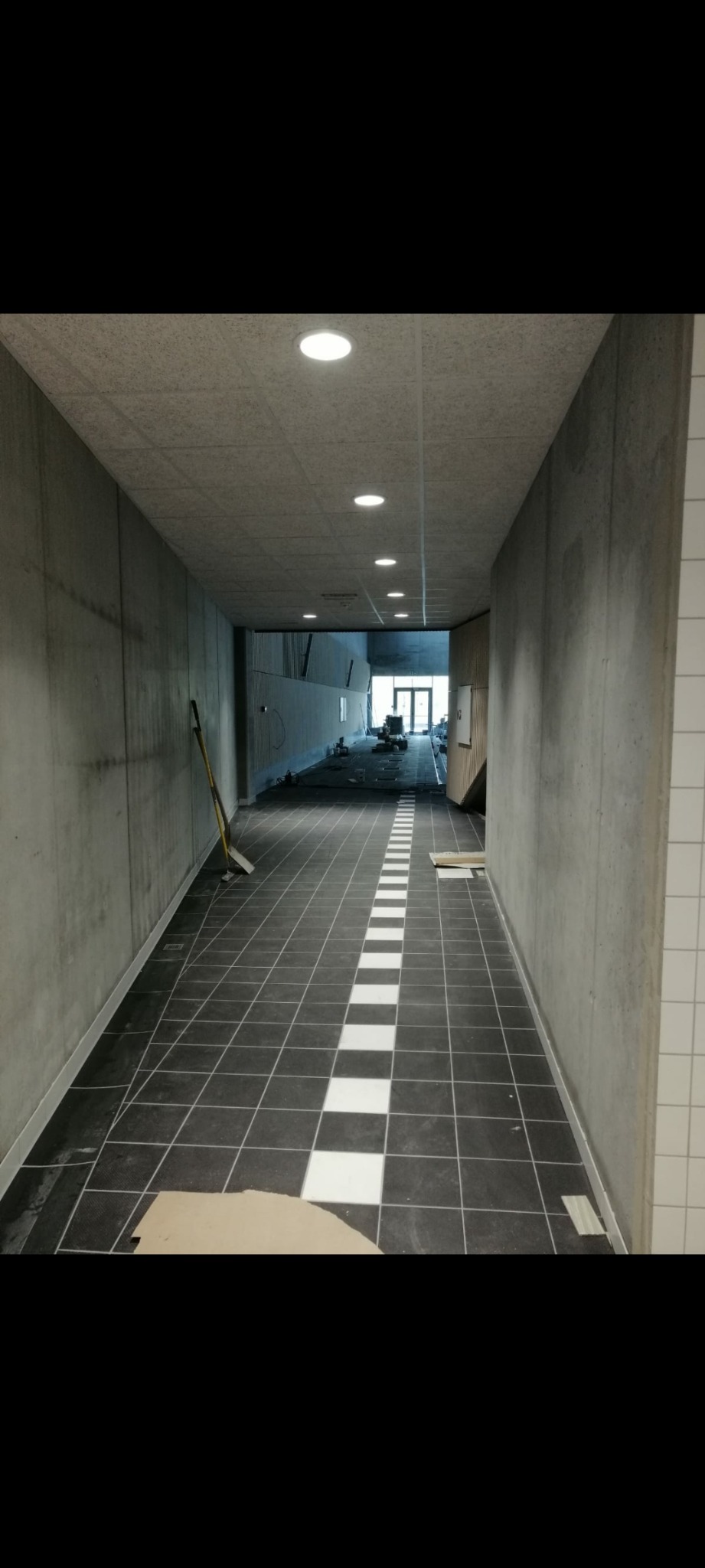 Korytarz w trakcie wykańczania, z czarno-białą szachownicą na podłodze, surowe betonowe ściany i widoczne narzędzia w tle.