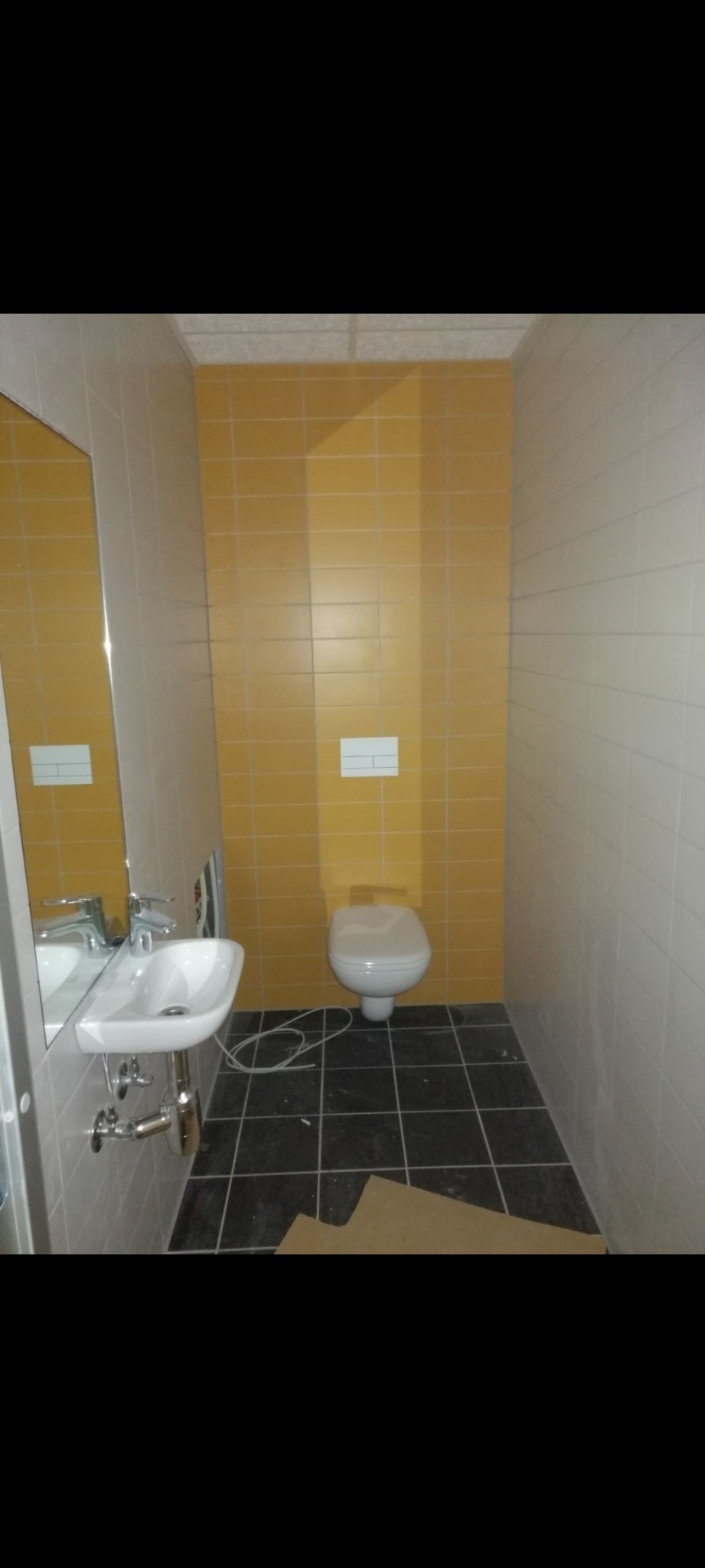Wnętrze łazienki w trakcie prac wykończeniowych: żółte i białe płytki na ścianach, czarna podłoga z resztkami kartonu, biała umywalka z lustrem i toaleta w tle.
