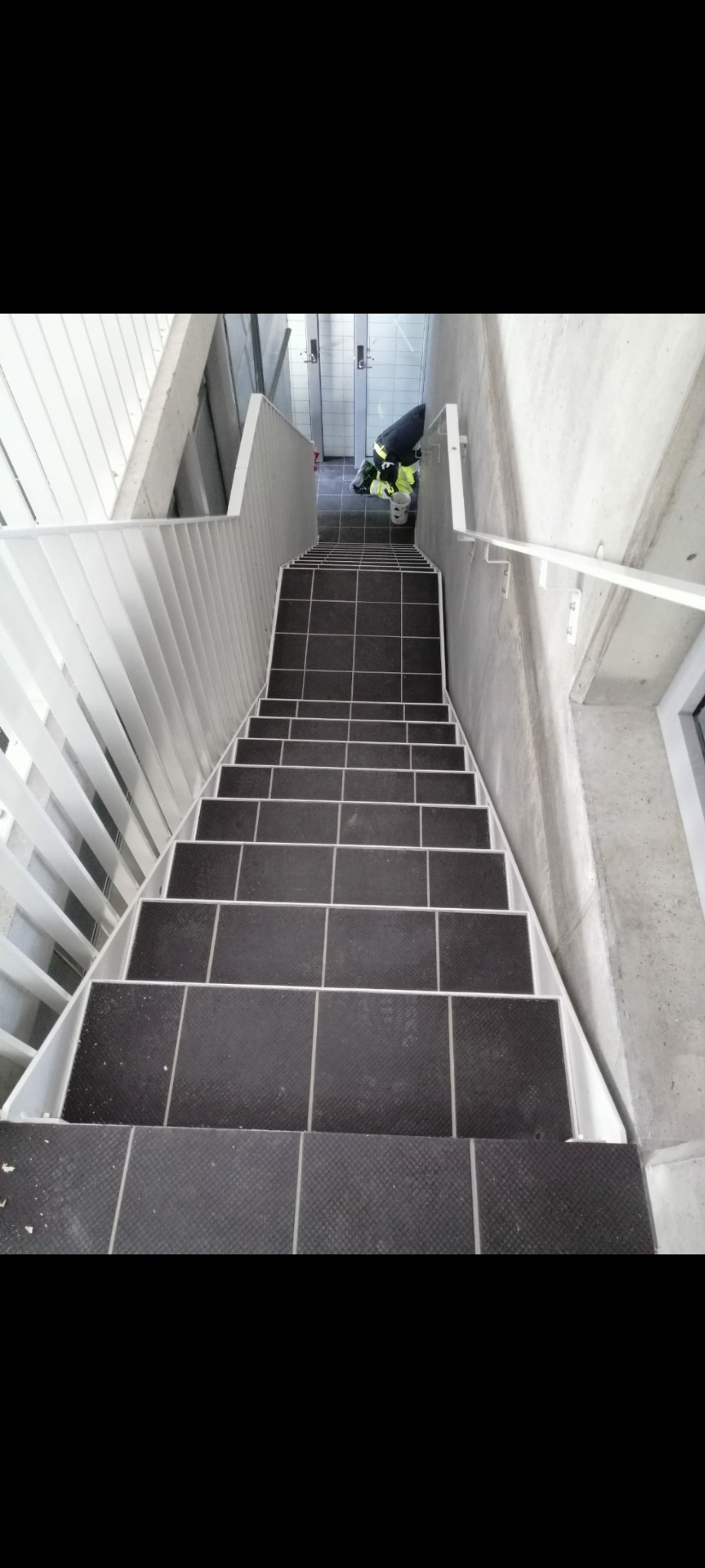 Schody wyłożone ciemnymi płytkami z antypoślizgową fakturą, białe barierki, widok z góry na klatkę schodową w budynku.