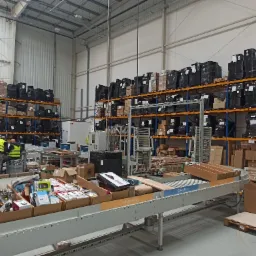 Wnętrze magazynu z regałami wypełnionymi zapakowanymi towarami, linia transportowa z różnymi produktami w kartonach, pracownicy w odblaskowych kamizelkach przy biurku.