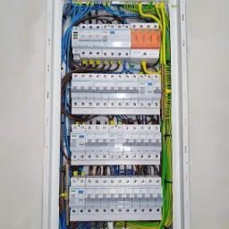 Rozdzielnica elektryczna z zainstalowanymi bezpiecznikami i przewodami w kolorach niebieskim, brązowym, żółto-zielonym oraz pomarańczowymi ogranicznikami przepięć, zamontowana w białej obudowie...