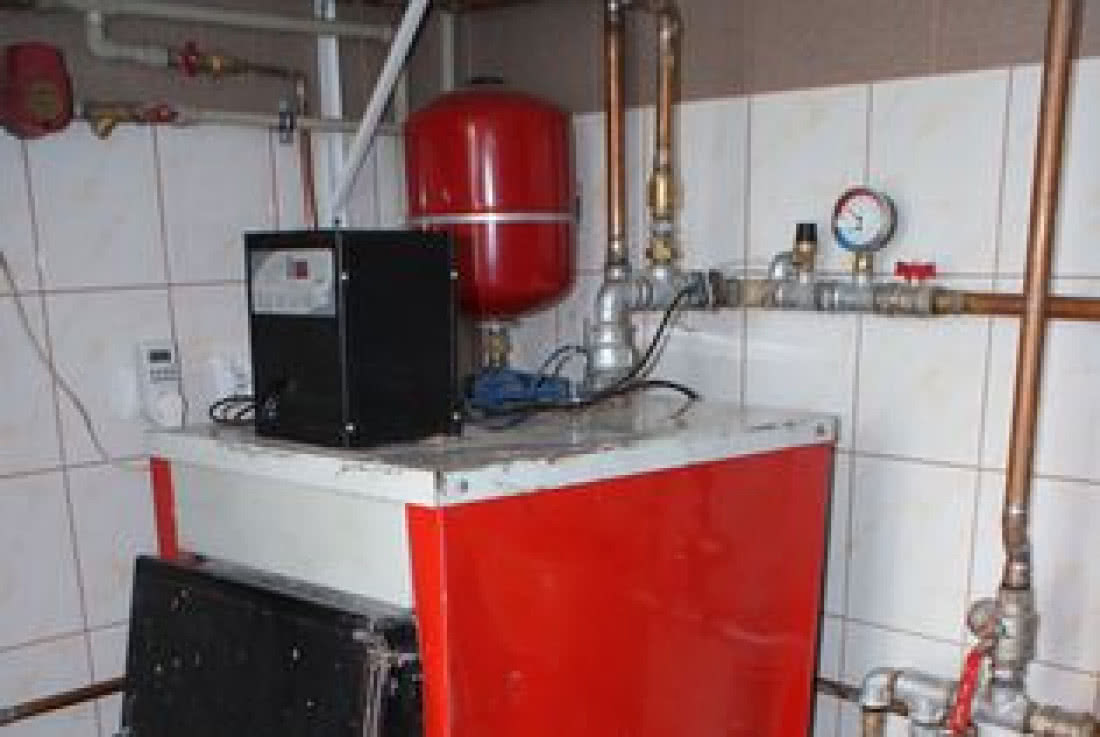 Czerwono-czarny piec grzewczy z podłączonymi miedzianymi rurami i manometrem na tle białych, kwadratowych płytek.