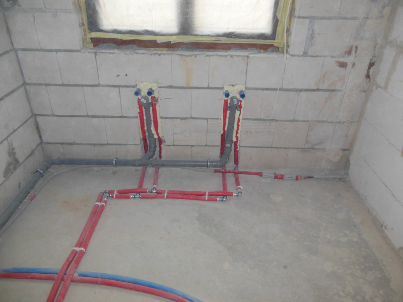 Surowa łazienka w trakcie remontu: widoczne nowe instalacje hydrauliczne z rur czerwonych i niebieskich, szare rury kanalizacyjne, ściany z białych bloczków, okno zabezpieczone folią.