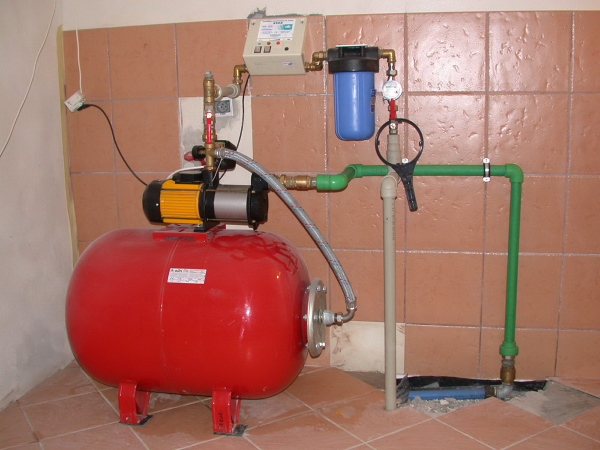 Czerwony hydrofor z żółto-czarnym silnikiem i zielonymi rurami instalacji wodnej w pomieszczeniu z brązowymi płytkami w Kaliszu. Widoczny filtr wody i manometr.