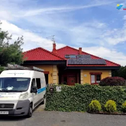 Żółty dom jednorodzinny z czerwoną dachówką i panelami słonecznymi na dachu, na tle błękitnego nieba z chmurami. Przed domem zaparkowany biały bus z logo firmy instalującej panele.