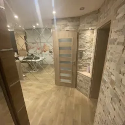 Korytarz z podłogą z paneli, ściana wyłożona kamieniem dekoracyjnym, drzwi wewnętrzne z przeszkleniami i ściana z lustrzanych płytek w romby odbijająca wnętrze pomieszczenia.