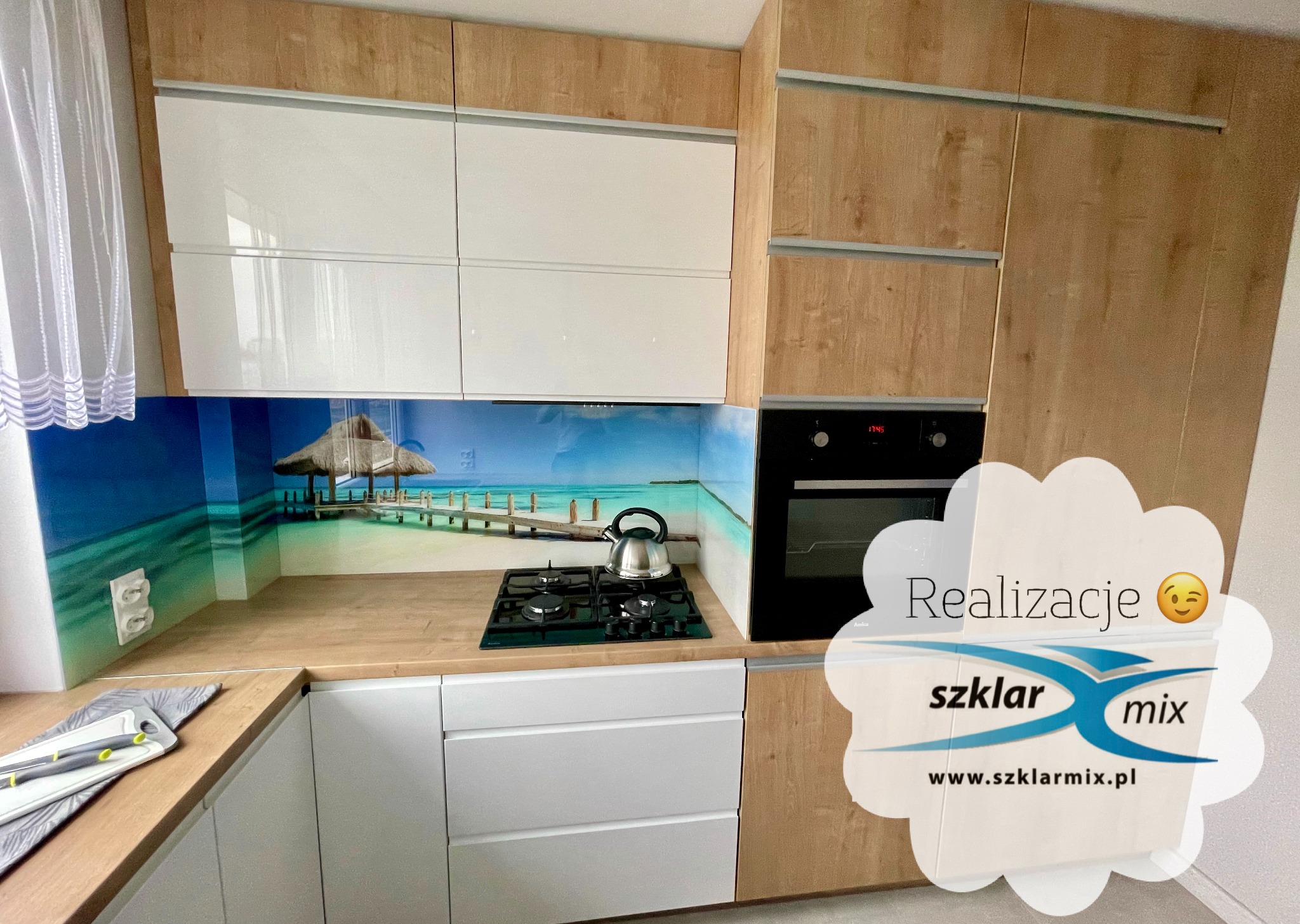 Kuchnia z panelami szklanymi z motywem plaży, zabudowa w kolorach drewna i bieli, czajnik na płycie grzewczej.
