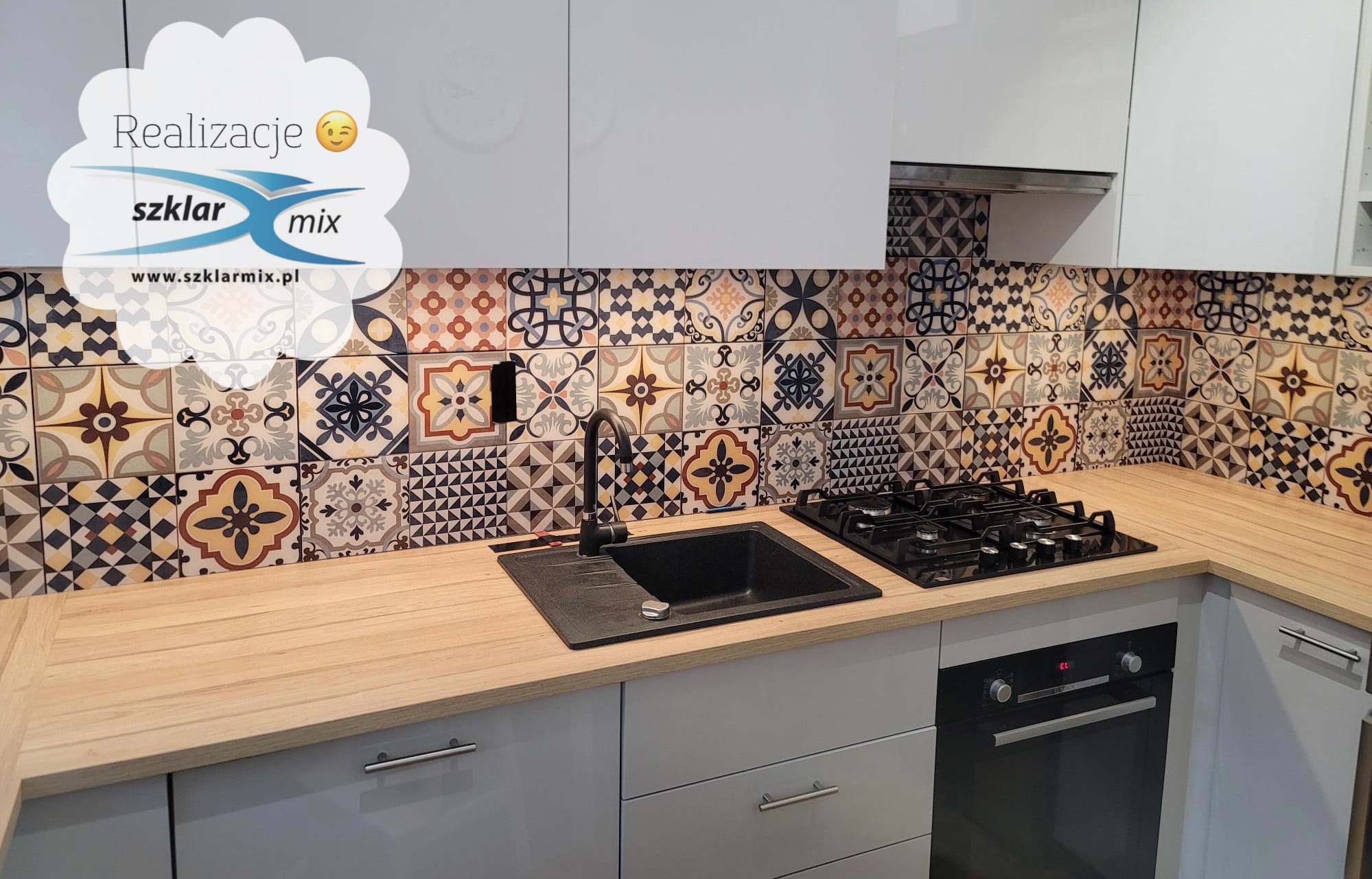 Kuchnia z panelami szklanymi z motywem patchworkowym nad blatem, czarny zlew i płyta gazowa, jasne szafki.