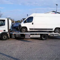 Biała laweta z napisem 'Pomoc Drogowa' i numerem telefonu przewozi białego dostawczaka na tle brukowanej nawierzchni i budynk&oacute;w.