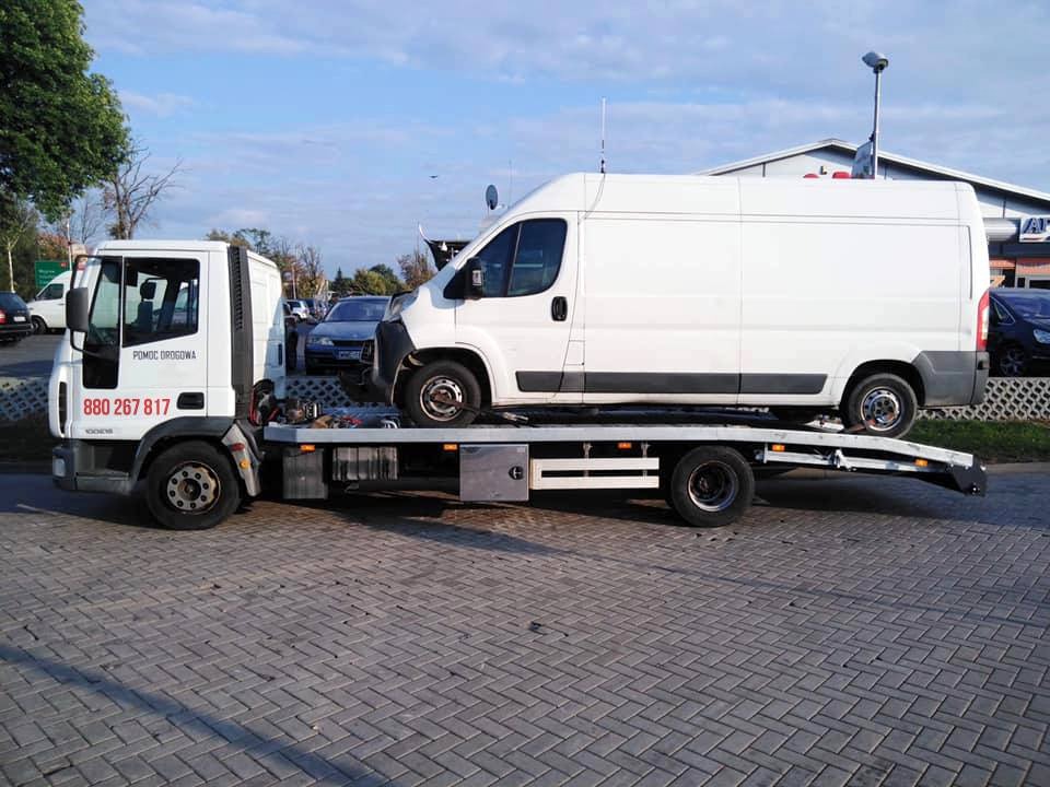 Biała laweta z napisem 'Pomoc Drogowa' i numerem telefonu przewozi białego dostawczaka na tle brukowanej nawierzchni i budynków.