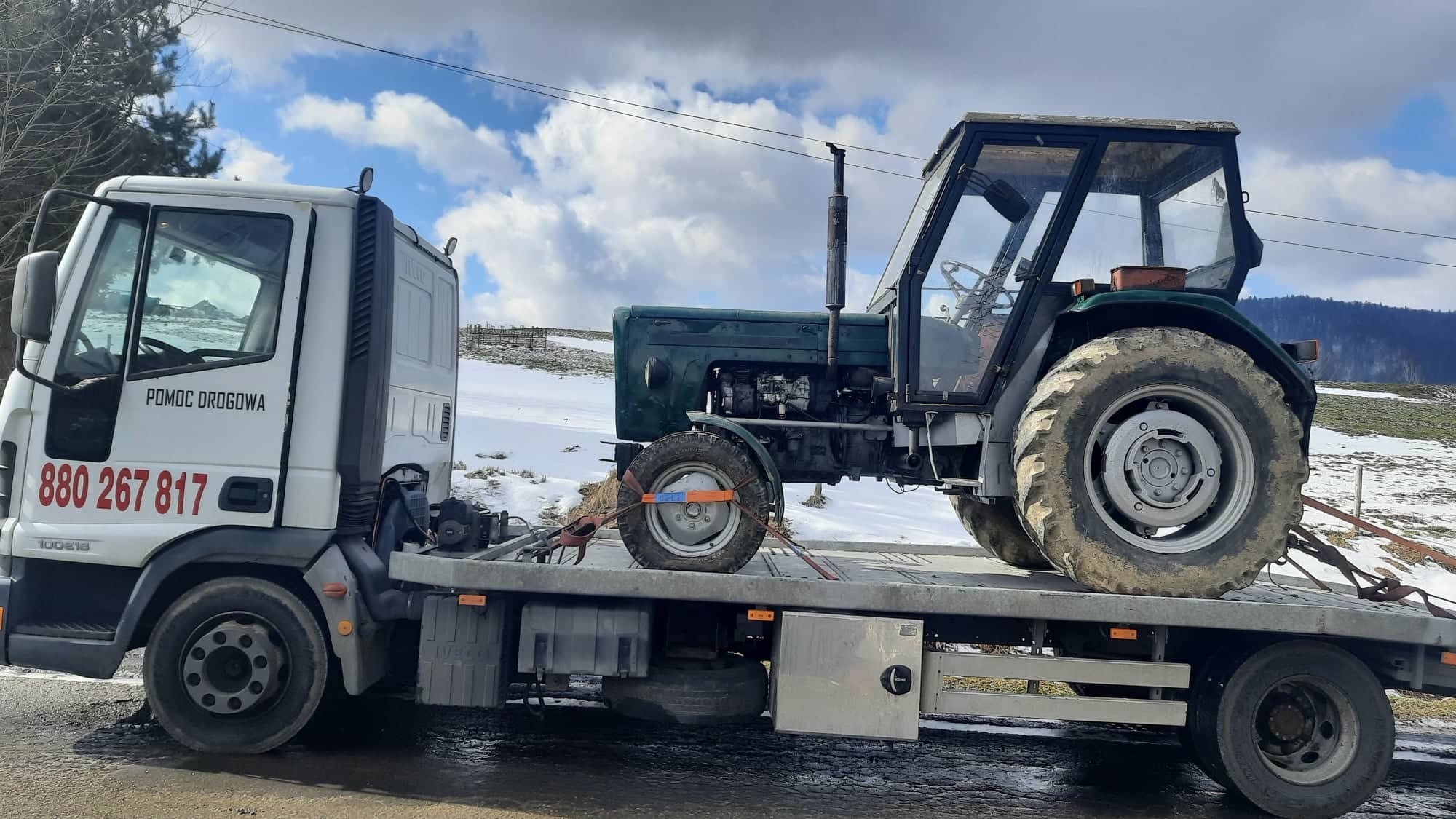 Zielony ciągnik rolniczy zabezpieczony pasami na platformie lawety pomocy drogowej z numerem telefonu na tle zimowego krajobrazu.