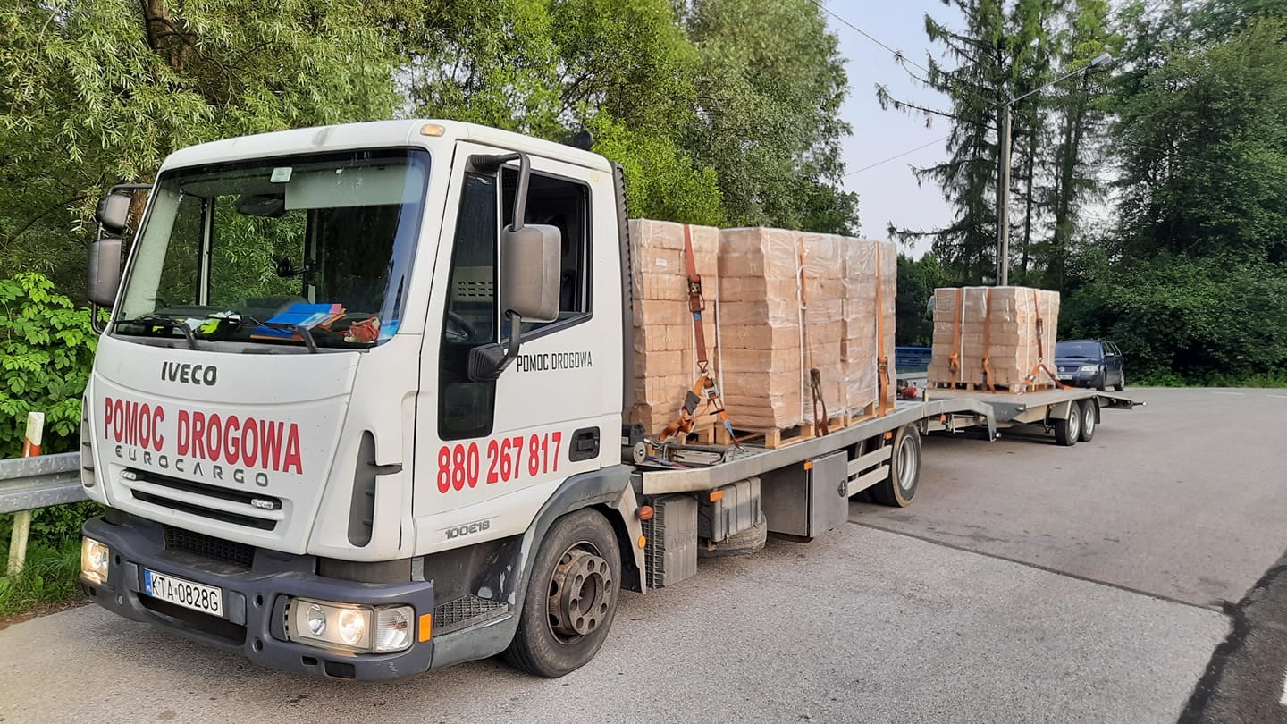 Biała laweta Iveco z napisem 'POMOC DROGOWA' przewozi zabezpieczone palety z cegłami na przyczepie, widok z boku na tle zieleni i asfaltowej drogi.