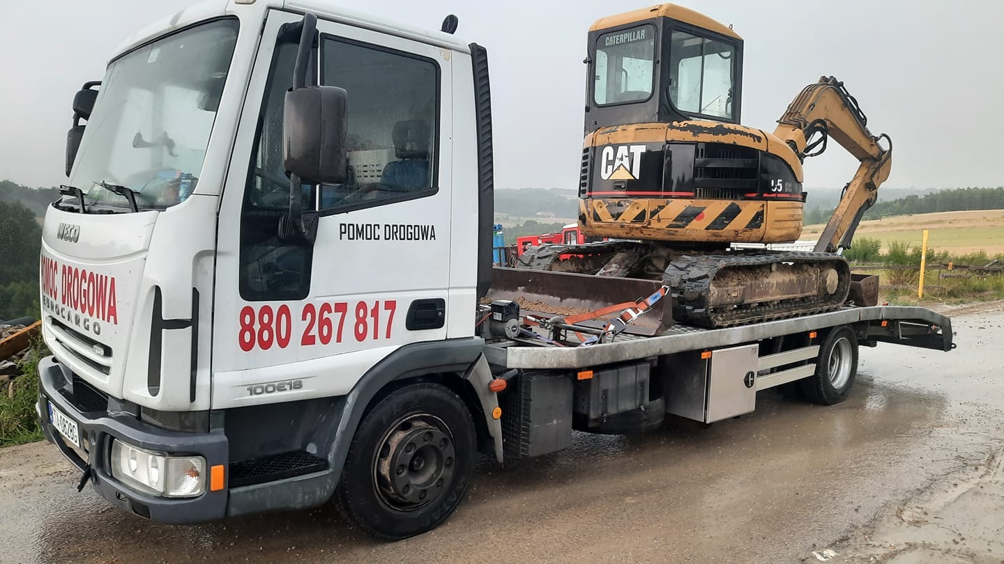 Biała laweta Iveco z napisem 'Pomoc Drogowa' przewozi żółtą, brudną koparkę Caterpillar na platformie, widok z boku na tle pola.